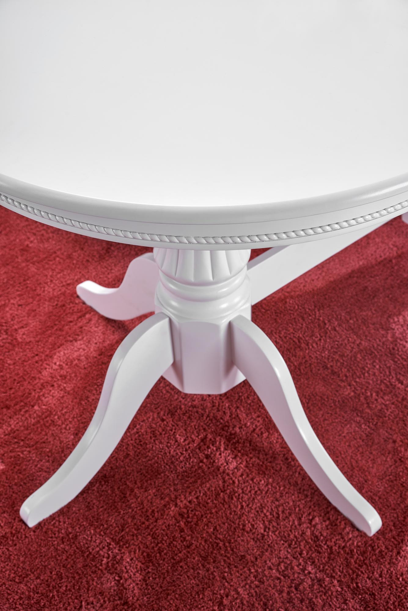 Zbliżenie na detal białego rozkładanego stołu JOSEPH z ozdobnym wykończeniem na czerwonym dywanie.
