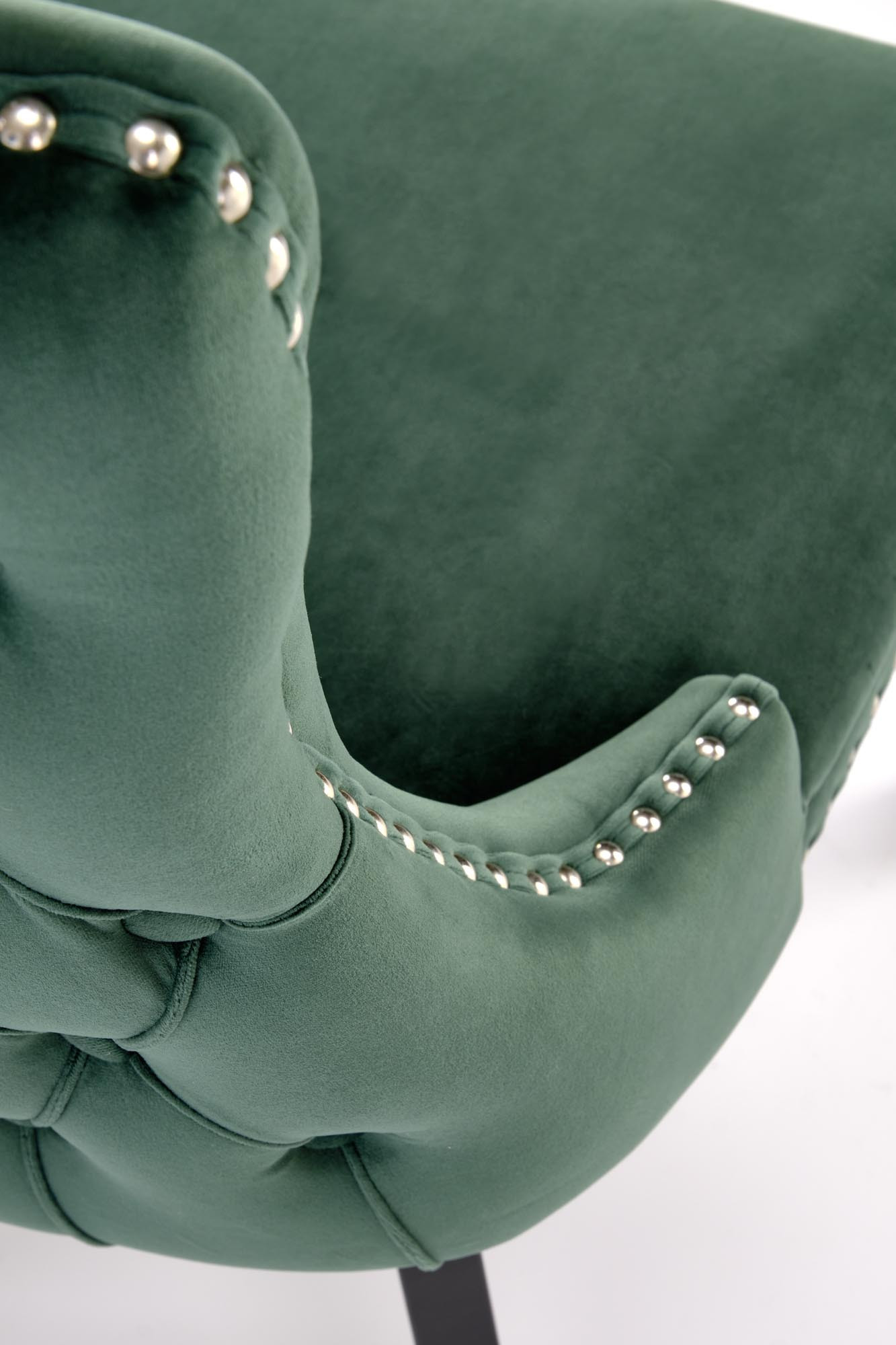 Zbliżenie na ciemnozielony aksamitny fotel ALDA z drewnianymi nogami i złotymi ćwiekami na bokach.